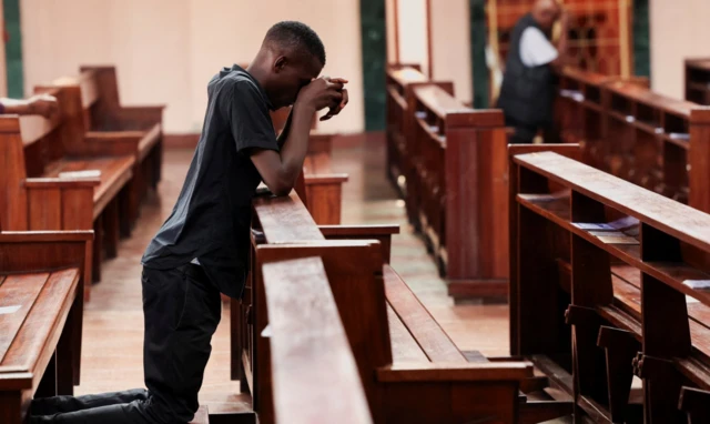 A Catholic faithful prays in a church. He is kneeling down and has bowed his head into his hands which are resting on wooden pews