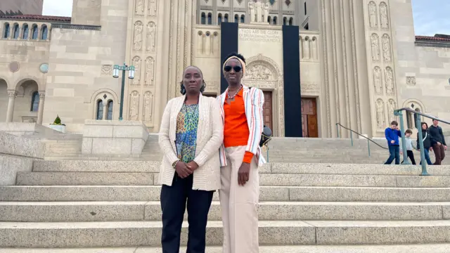 Two women pose for a photo. On the left, Nina, is wearing a cream-coloured sweater over a a mosiac blouse and a black rosary. On the right, Landrada is wearing an orange blouse and a stripped sweater. She is wearing black sunglasses and a headband.