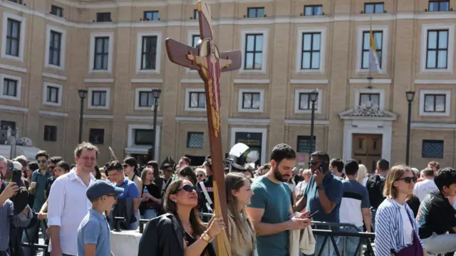 A woman is carrying a large cross as she stands in a crowd