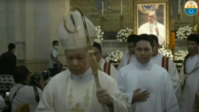 Screenshot of a livestream, showing a priest and other members of the clergy walking in a line away from a framed photo of Pope Francis