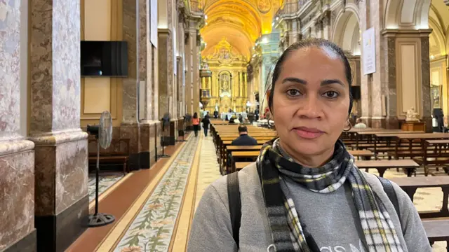 A woman standing in a cathedral looks at the camera.