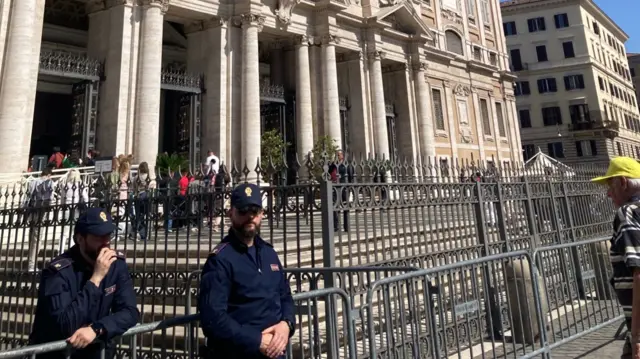 Two Italian police officers (L) stand guard outside a church in their blue uniforms and matching caps. To their left, a man in a yellow cap and striped shirt observes the metal barriers