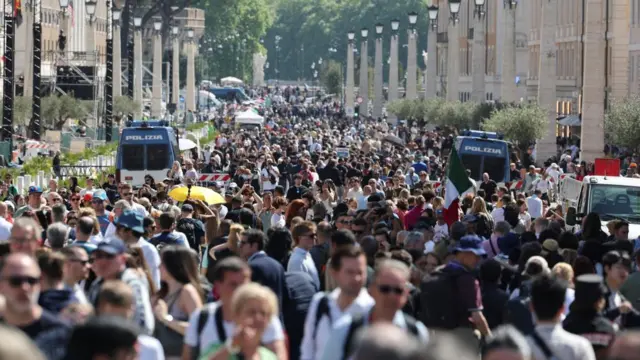 A large crowd in the vicinity of St Peter's Basilica. Two blue police vans are also situated on the street