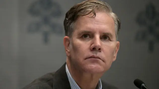 Juan Carlos Cruz Chellow looking serious, wearing a suit and with a microphone in front of him