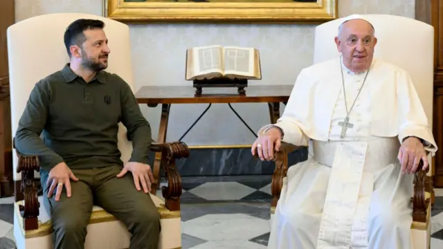 Volodymyr Zelensky (L) sitting on a cream-white chair in green long-sleeved polo and trousers. Pope Francis (R) sits in a matching chair in his white cassock and skullcap