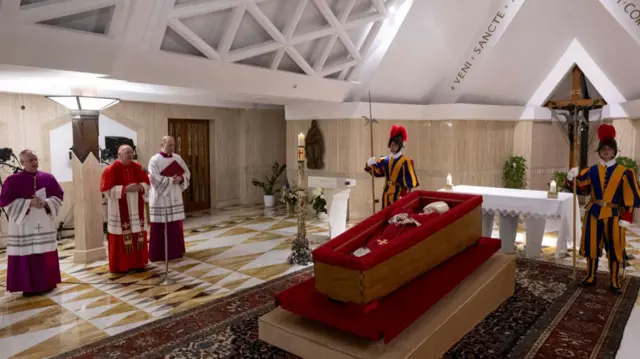 The remains of Pope Francis lay inside a wooden casket lined with red cloth and resting on top of a wooden catafalque as two Swiss Guards stand to its sides