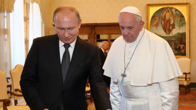 Vladimir Putin (L) stands next to Pope Francis (R) as they observe some objects laying on a table off frame