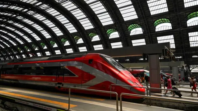 A red train waits at the platform at a train station in Italy as passengers walk along with their bags