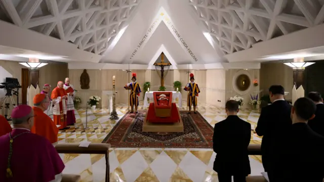 The Pope lays in a wooden coffin dressed in red, with rosary beads draped across his hands, and guards either side