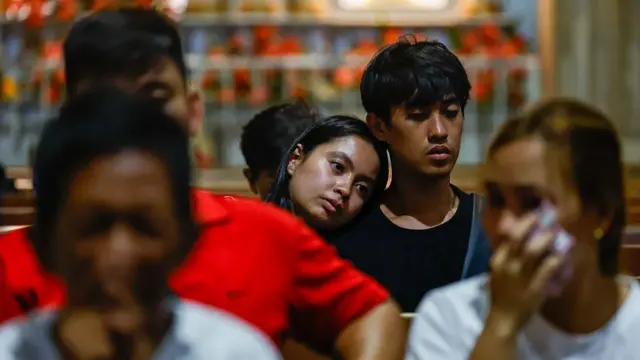 A young woman leaning on the shoulder of a young man, both seated on church benches. In front of them are older women dabbing their faces with handkerchiefs or tissue paper