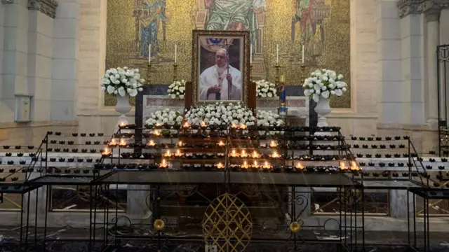 An altar of with a rack of lit candles and a large framed photo of Pope Francis