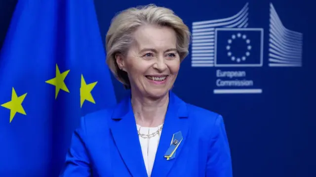 EU Commission President Ursula von der Leyen standing in front of a blue wall and an European flag. She's wearing a blue suit with a white blouse and a pin in the colours of Ukraine on his left lapel