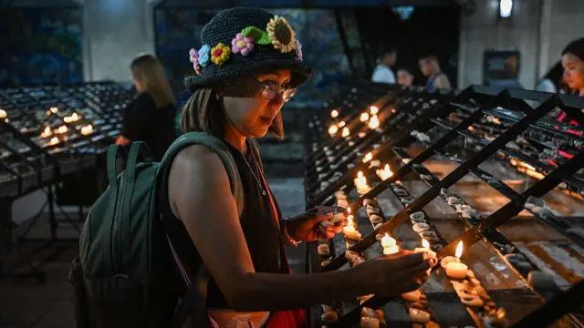 A woman wearing a colourful hat and backpack placing a lit candle among other candles on a rack