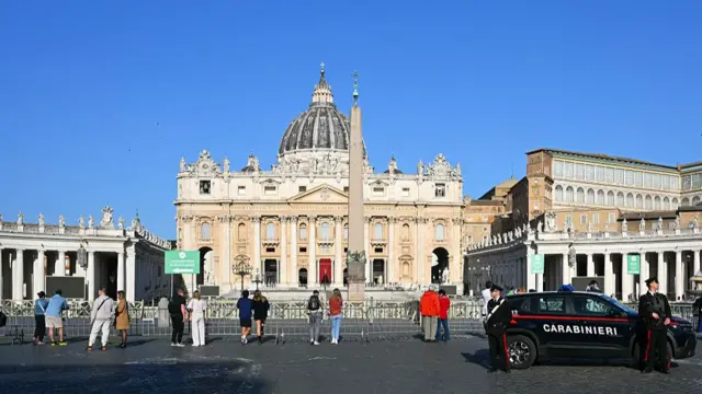 St Peter's Basilica exterior mid-morning as some tourists walk around the front square. A Carabiniari military police car with two officers on the right side of the frame