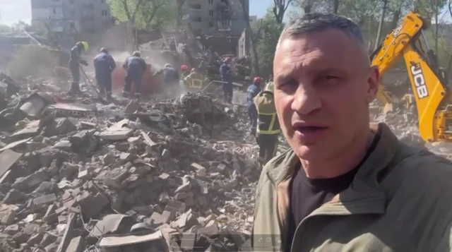 Vitali Klitschko speaking into a camera in front of a Russian strike site. Emergency workers are digging through the rubble and using a JCB excavator. Smoke is rising from parts of the rubble.