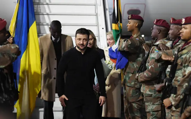 Zelensky walks towards the camera, military personnel in uniform stand to his left and right