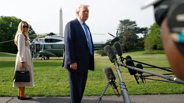 Trump stands in a suit and blue tie, speaking in front of several microphones. Melania Trump wears a brown trench coat and black sunglasses and a black bag, standing behind Trump. Marine One helicopter is in the background.