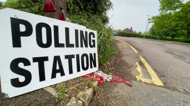 A sign on the side of the road that says Polling Station in black lettering against a white background.