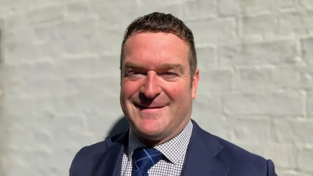 Profile picture of Ryan Coogan, he wears a navy suit jacket, white checked shirt and navy tie with stripes. The background is a brick wall that has been painted white.