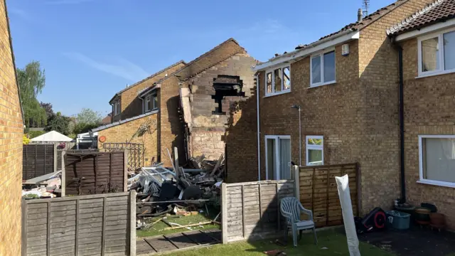 A view across the neighbouring back garden towards the destroyed home. It shows charred bricks inside the home and piles of debris on the ground.