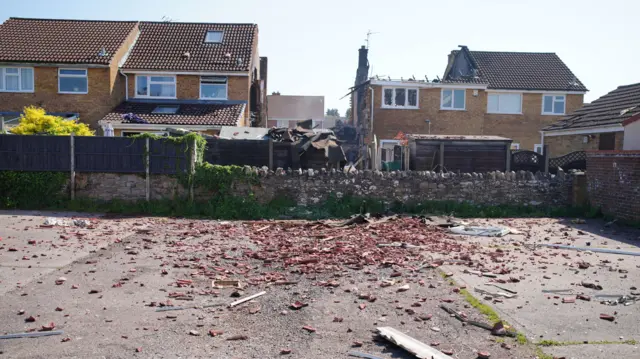 A view over the back garden of the damaged property, showing the row of terraced homes with the middle one burnt to the ground. In the foreground, the car park is covered in brick fragments and debris.