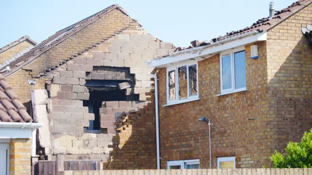 The charred bricks of the damaged home. The roof has completely gone but the line where the tiles once were is still visible. A neighbour's bedroom window has been smashed from the impact.