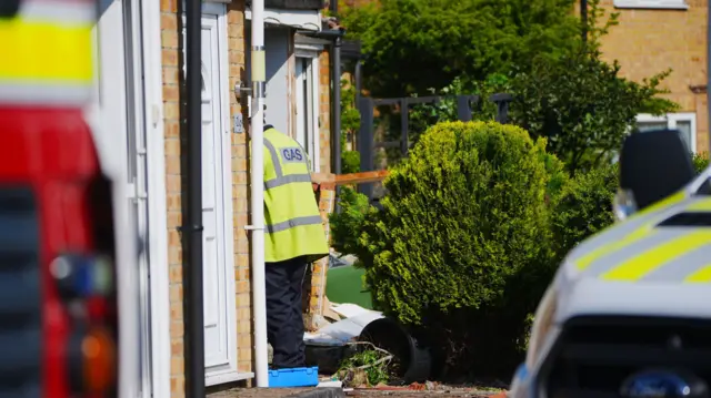 A person wearing black trousers and a yellow hi-vis jacket that says 'GAS' across the back. They are entering the front door of a property. Emergency vehicles are visible in the foreground, out of focus.