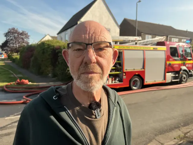 A man standing in front of a fire engine on a street. He is wearing a green hoodie, brown top, glasses and is looking at the camera