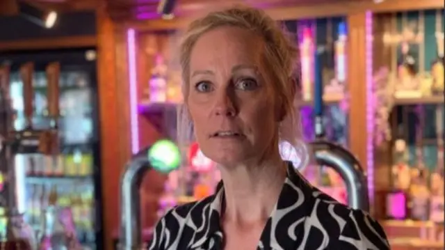 A woman with blonde hair looking to the camera. She has on a black and white shirt and is stood in a pub.
