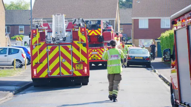 A female fire fighter wearing a yellow hi-vis jacket and khaki trousers. She is signalling to two fire engines who are reversing up Lancaster Road