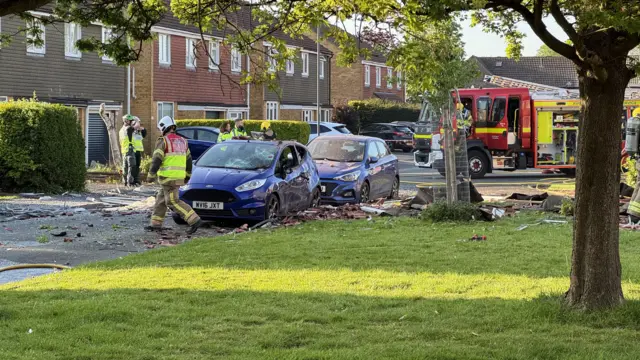 Parked cars on Lancaster Road that have been damaged by the impact from the explosion and falling debris. Firefighters are standing outside the property and there is a fire engine parked on the right.