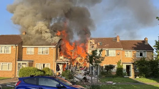 A row of terraced brick houses. The one in the middle has been completely gutted by fire, with only part of the front porch remaining. The neighbouring homes have sustained fire damage to their roofs. There are bright orange flames actively burning, and a large plume of black smoke billowing into the sky.