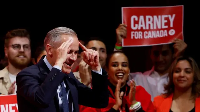 Mark Carney smiling and pointing at his own head, while supporters in the background smile and clap