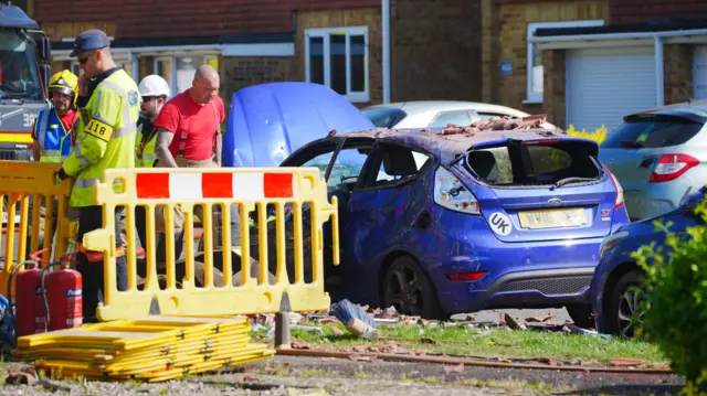 A blue Ford parked on the road at the scene of the explosion. Its roof has been caved in from falling bricks and debris. Emergency crews have set up