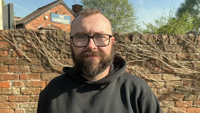 Rich Cainey wearing a black hoodie and black rectangular glasses. He has a full dark beard and short brown hair which is shaven on the sides. He sis standing in front of a brick wall outside the Farmhouse pub.