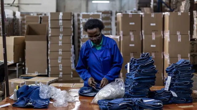 A worker making pairs of jeans in a factory in Lesotho. She wears blue overalls, behind her is a large pile of cardboard boxes and to her right, two piles of blue denim jeans.
