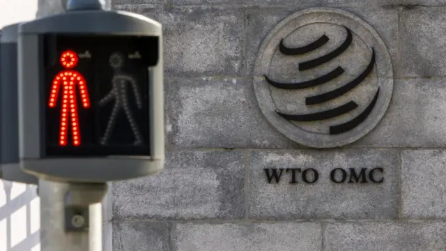 The exterior of the WTO - its logo is on a stone wall. To the left, a red pedestrian crossing signal shows