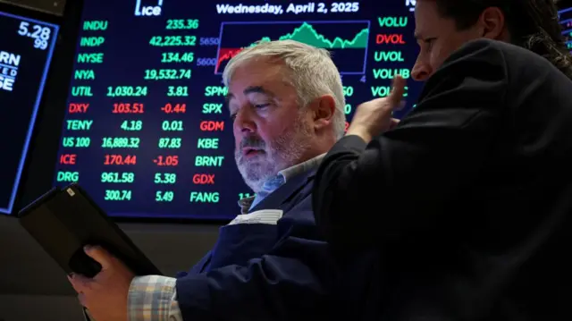 Traders work on the floor at the New York Stock Exchange, with a screen showing charts and numbers in the background.