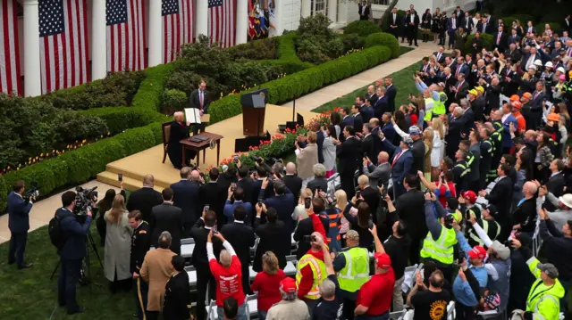 Donald Trump holds up his executive order at a desk in the White Hose Rose Garden, in front of a large audience