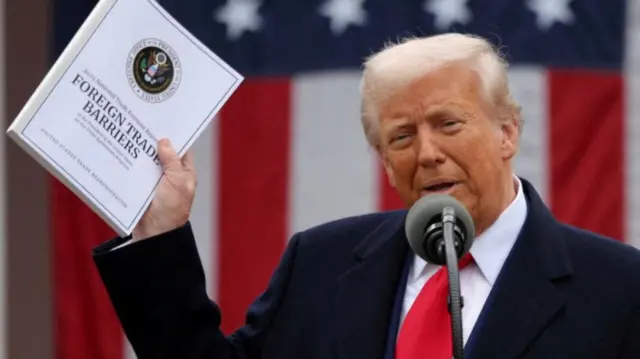 Trump standing speaking into a big microphone. He holds a thick white booklet with the words foreign trade barriers on it. Behind him is a large US flag
