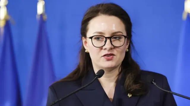 Yulia Svyrydenko in a black blazer delivers speech while standing in front of a series of European Union flags and a blue background
