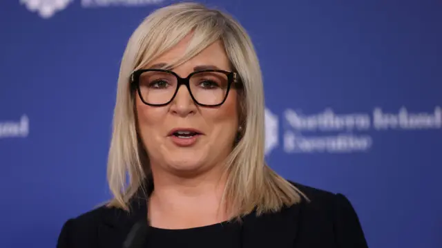 Michelle O’Neill speaking at a press conference. She has blonde hair and brown eyes, and is wearing black-rimmed glasses.