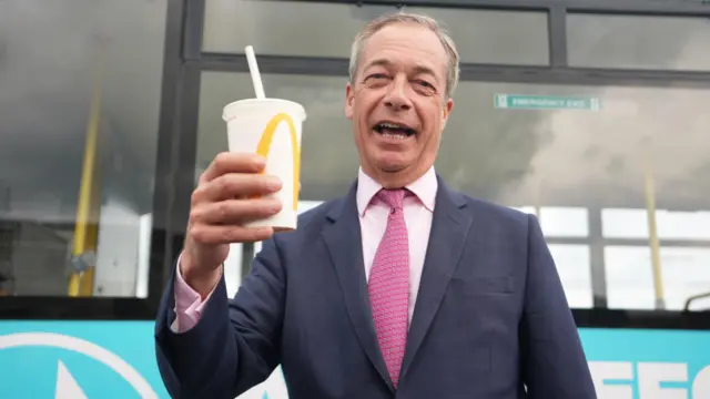 Nigel Farage holding a MacDonald's milkshake in his right hand. He is wearing a jacket with a pink shirt and tie.