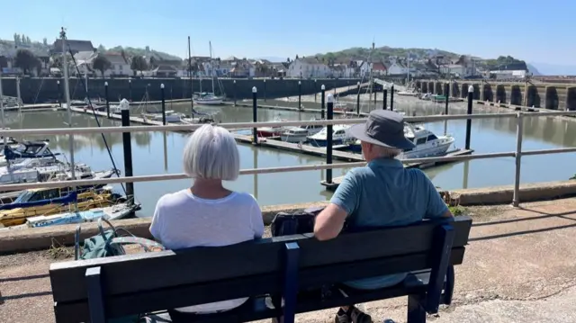 Two people sitting in a bench and looking out across a small harbour and coastal village
