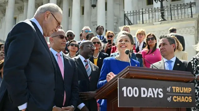Chuck Schumer and Democratic congressmembers stand in front of the Capitol in front of a podium that says "100 days of Trump chaos"