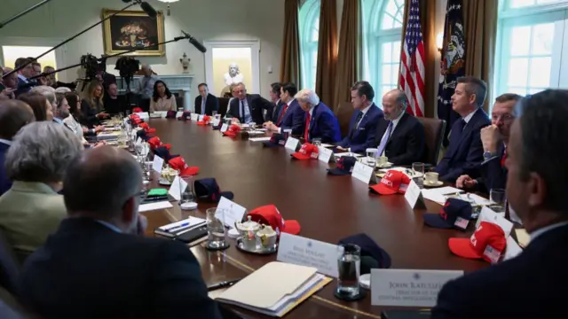 A table with lots of people sat around it including the president in the middle. Stood on one side is a row of journalists holding long microphones over the table