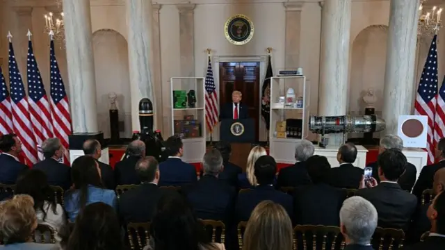 Trump speaking in a room full of rows of people sitting watching him. The room is fancy looking with marble columns and several US flags on stands either side of him