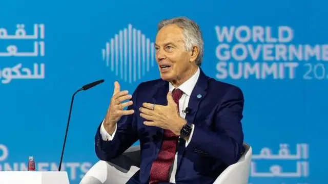 Tony Blair in a navy suit and red tie gestures with his hands while speaking at the World Government Summit