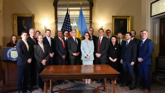 A room full of diplomats in suits with a table in the middle, and the US and Ukraine flags.