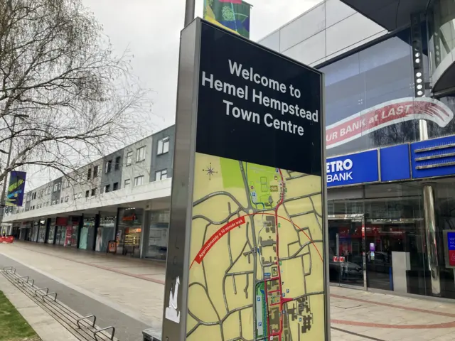 A picture of a sign in Hemel Hempstead that says "Welcome to Hemel Hempstead Town Centre" on it. Behind it is a row of shops that are part of the Marlowes shopping centre
